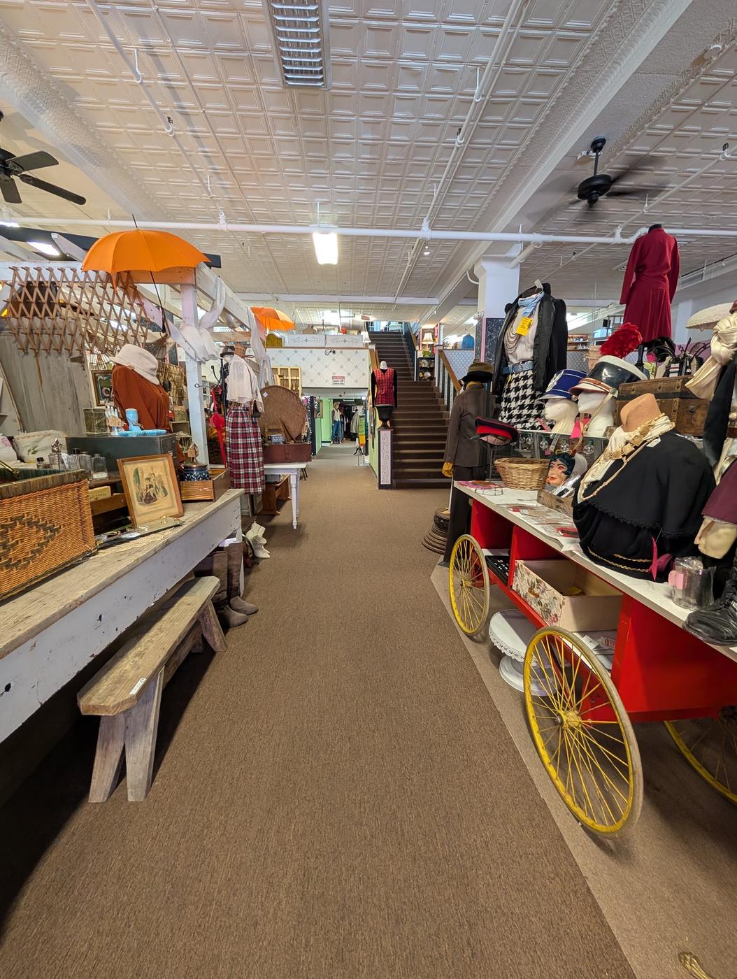 An aisle at Nostalgia with a red antique cart on yellow spoked metal wheels, mannequins in hats and houndstooth, orange parasols overhead and a staircase climbing out of frame.